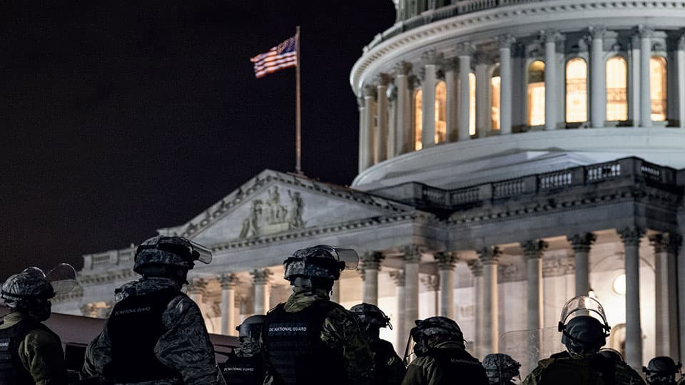 The image is from Elesa Zehndorfer's book Post-Truth Politics: A Brave New World?. There are eight people dress in army outfits outside a building with an american flag on it.